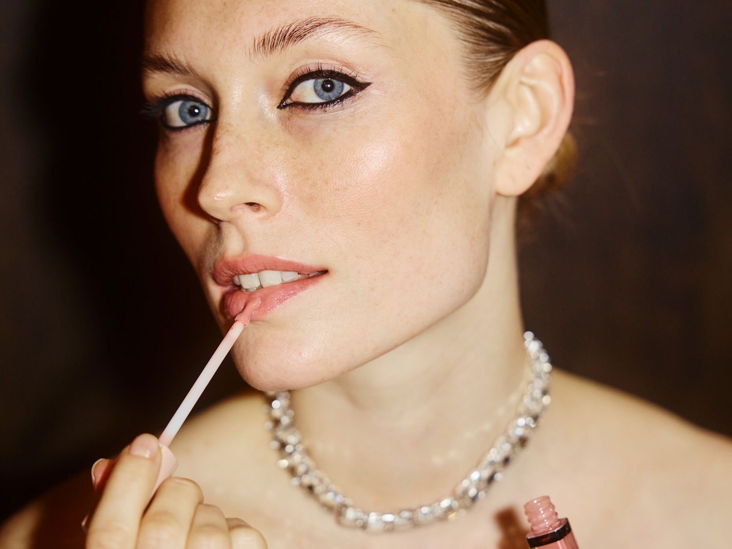 Een vrouw met blauwe ogen en een zilveren ketting brengt een roze lipgloss aan voor een subtiele glans.