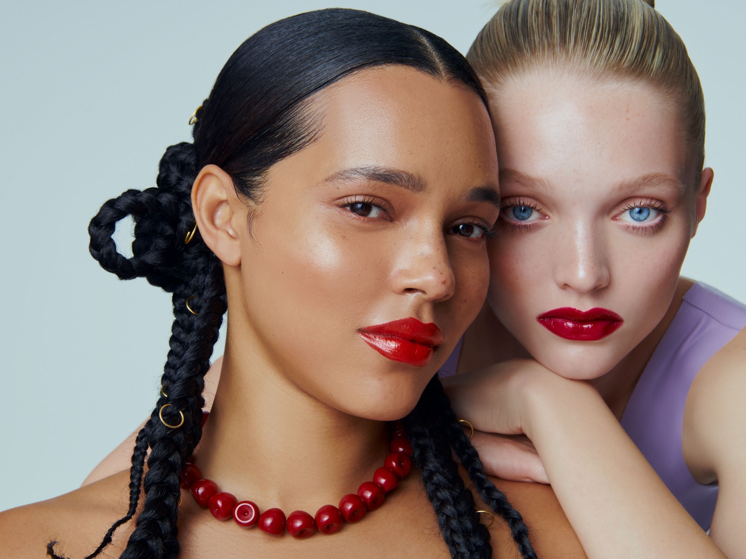 Nahaufnahme von zwei Models mit glänzenden roten Lippen, wobei das eine dunkle Zöpfe und eine rote Halskette trägt und das andere hellhäutig mit blonden Haaren ist.