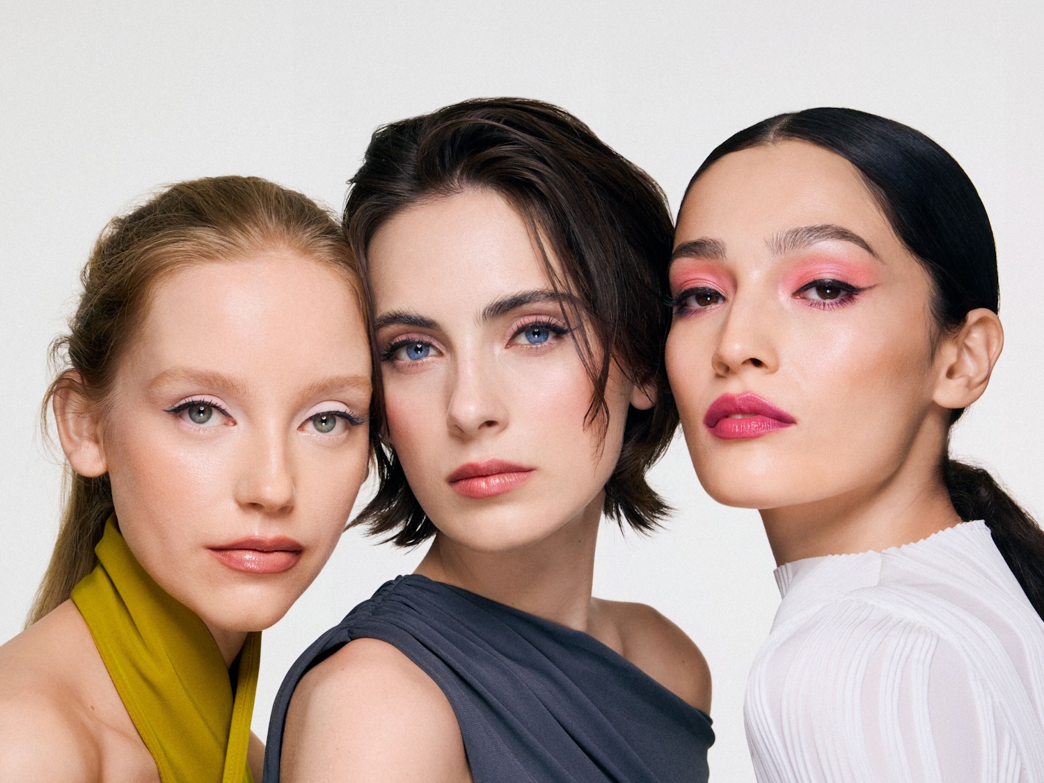Drei Frauen mit unterschiedlichen Hauttönen und Haarfarben präsentieren auffällige Make-up-Looks, darunter weißen Eyeliner, blauen Lidschatten und leuchtend pinke Lippen.