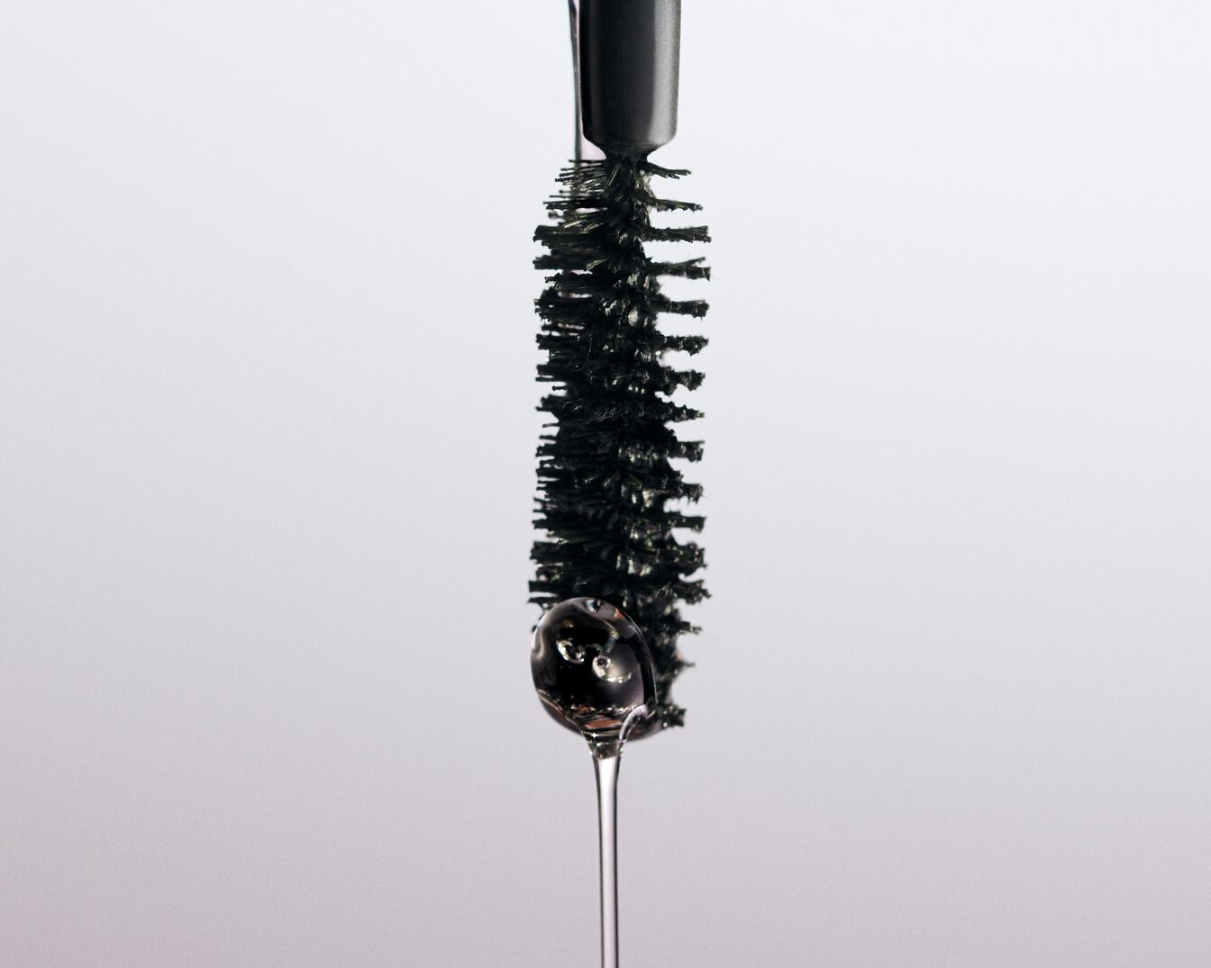 Close-up van een mascara borstel met een druppel zwarte vloeistof die eraf valt, tegen een witte achtergrond.