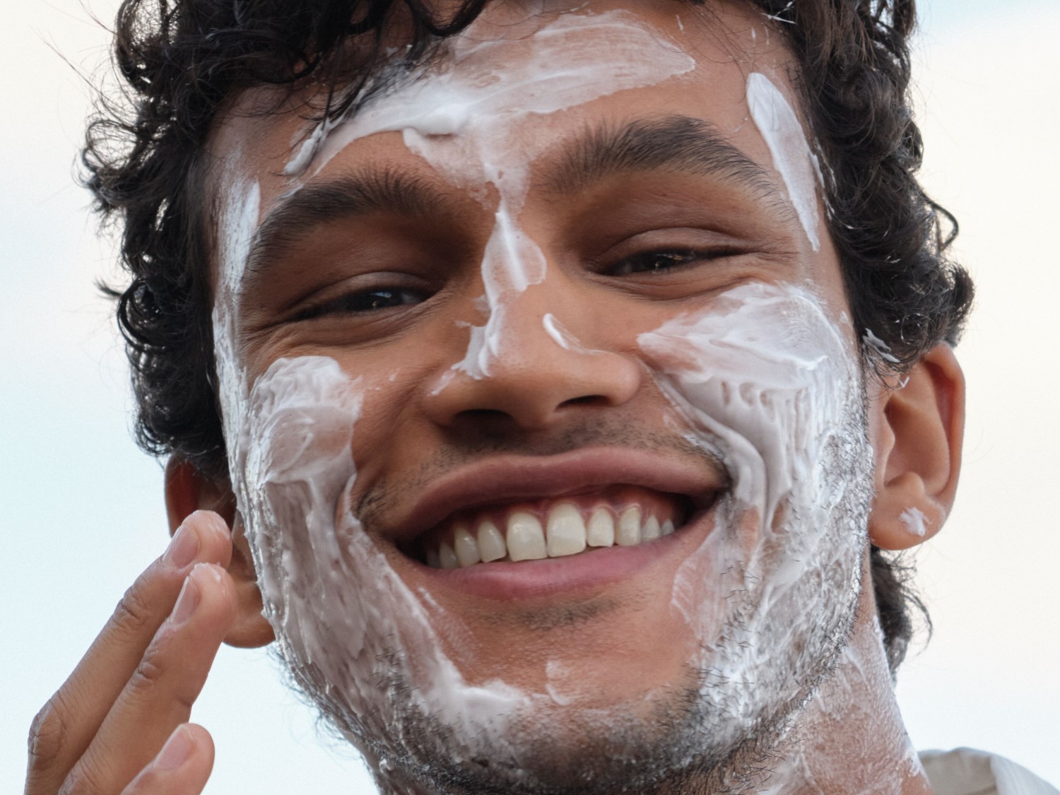Een lachende man met krullend haar smeert witte zonnebrandcrème op zijn gezicht, waarbij hij zijn huid beschermt tegen de zon.