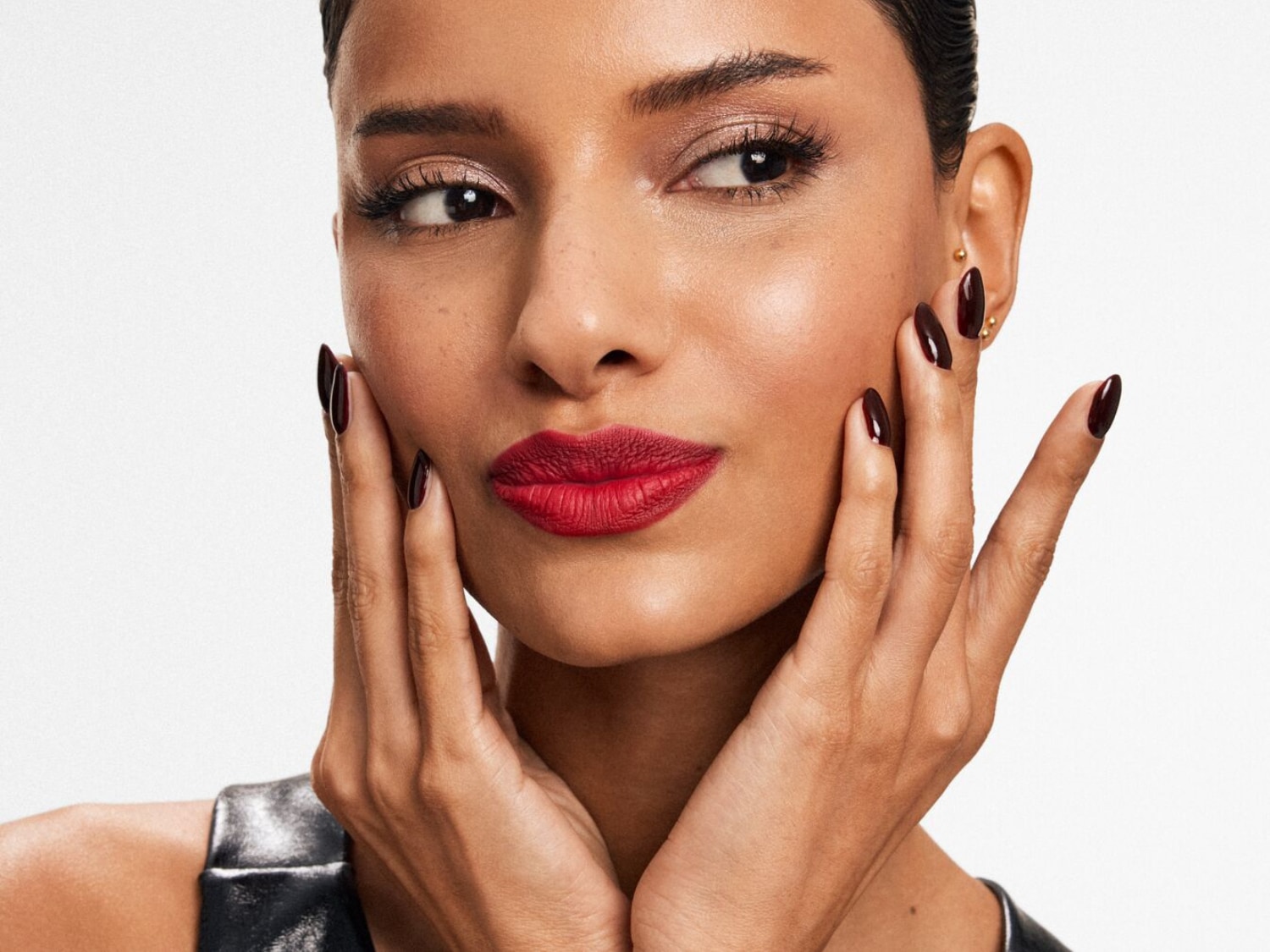 Portret kobiety z ciemnymi włosami, czerwonymi ustami i ciemnymi paznokciami, która dotyka dłońmi policzka, prezentując makijaż i manicure.