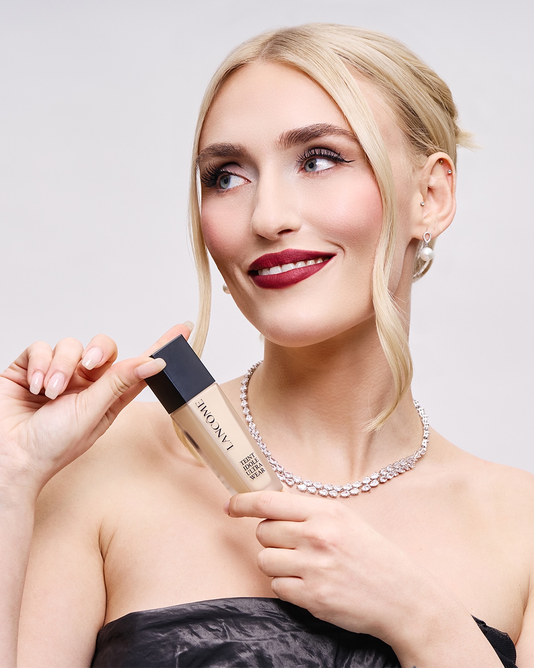 Una donna bionda, con un elegante abito nero e una collana di diamanti, tiene in mano il fondotinta Lancôme Teint Idole Ultra Wear, esaltandone la texture e la tonalità.