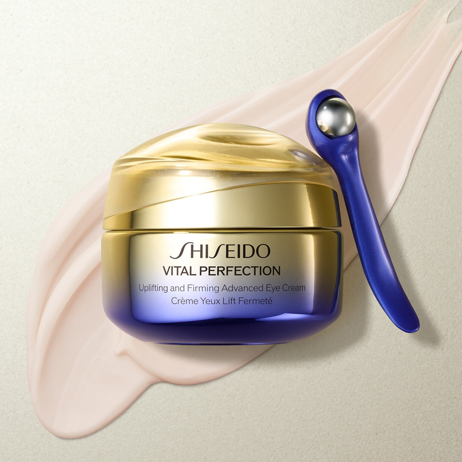 Ecco il vasetto dorato di Shiseido Vital Perfection, una crema occhi liftante e rassodante, con applicatore blu, su una pennellata di crema beige.