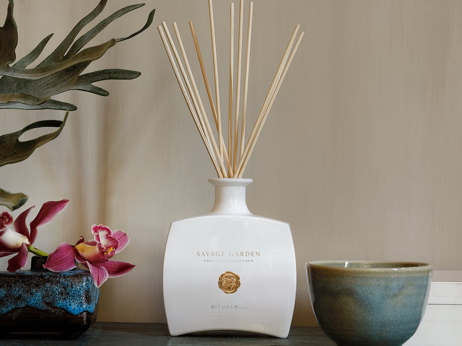 A Rituals Savage Garden illatdiffúzor fehér üvegben, fa pálcikákkal, virágokkal és kerámia edényekkel körülvéve.