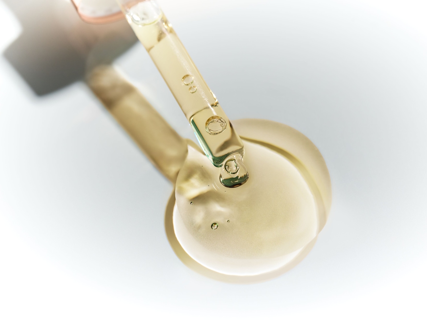Ein goldfarbenes Serum wird mit einer Pipette auf eine glatte, weiße Oberfläche aufgetragen, wobei sich ein kleiner Tropfen des Serums auf der Oberfläche sammelt.