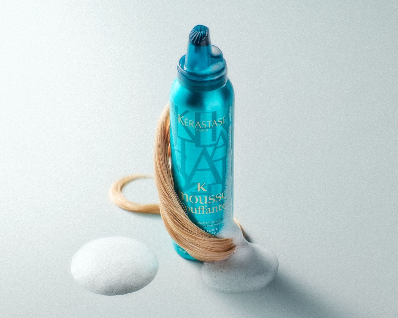 Türkisblaue Dose Kérastase K Mousse Bouffante mit einer Strähne blonden Haares und einem Klecks Schaum, der Volumen und Halt verspricht.