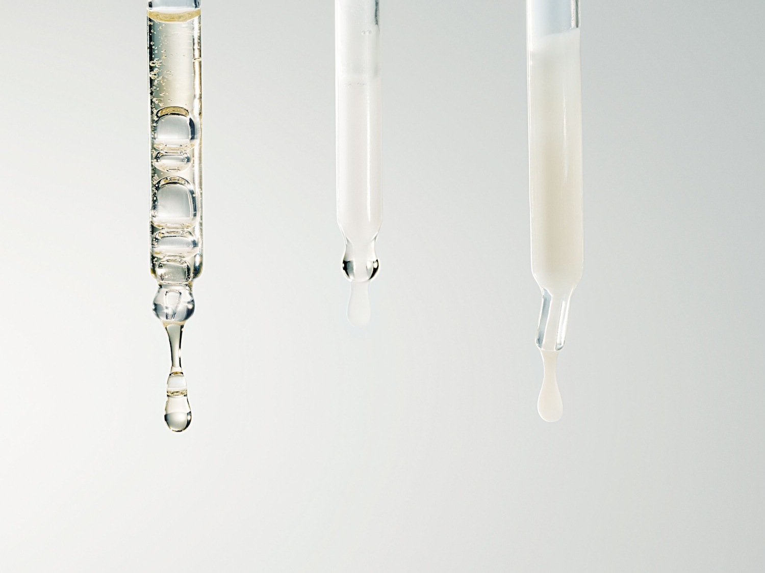 Drei Pipetten mit verschiedenen Texturen: eine klare Flüssigkeit mit Luftblasen, eine milchig-weiße und eine cremefarbene Flüssigkeit.