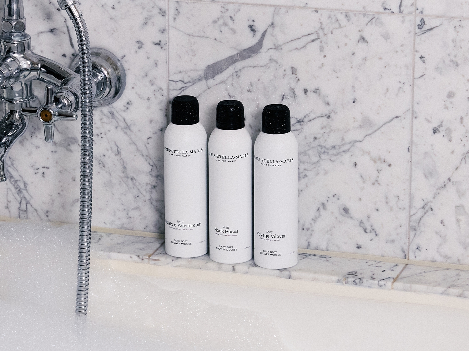 Drie witte flessen Marie-Stella-Maris silky soft shower mousse staan op de rand van een marmeren badkuip, met de geuren Objets d'Amsterdam, Rock Roses en Voyage Vétiver.