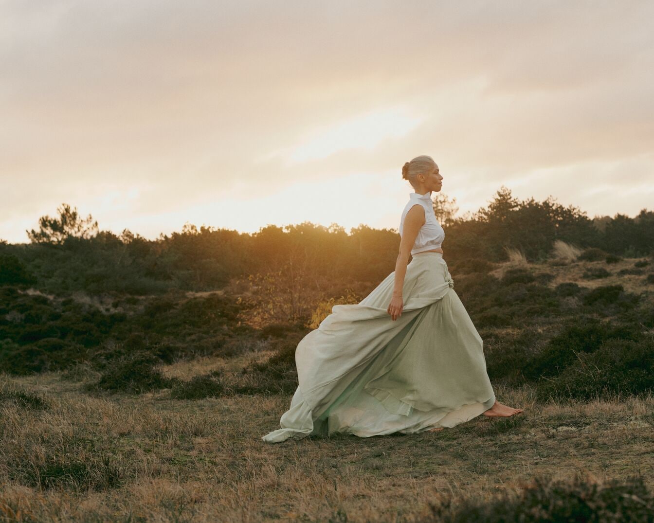 Immagine di una donna in un campo al tramonto, che indossa un vestito lungo e che cammina verso destra