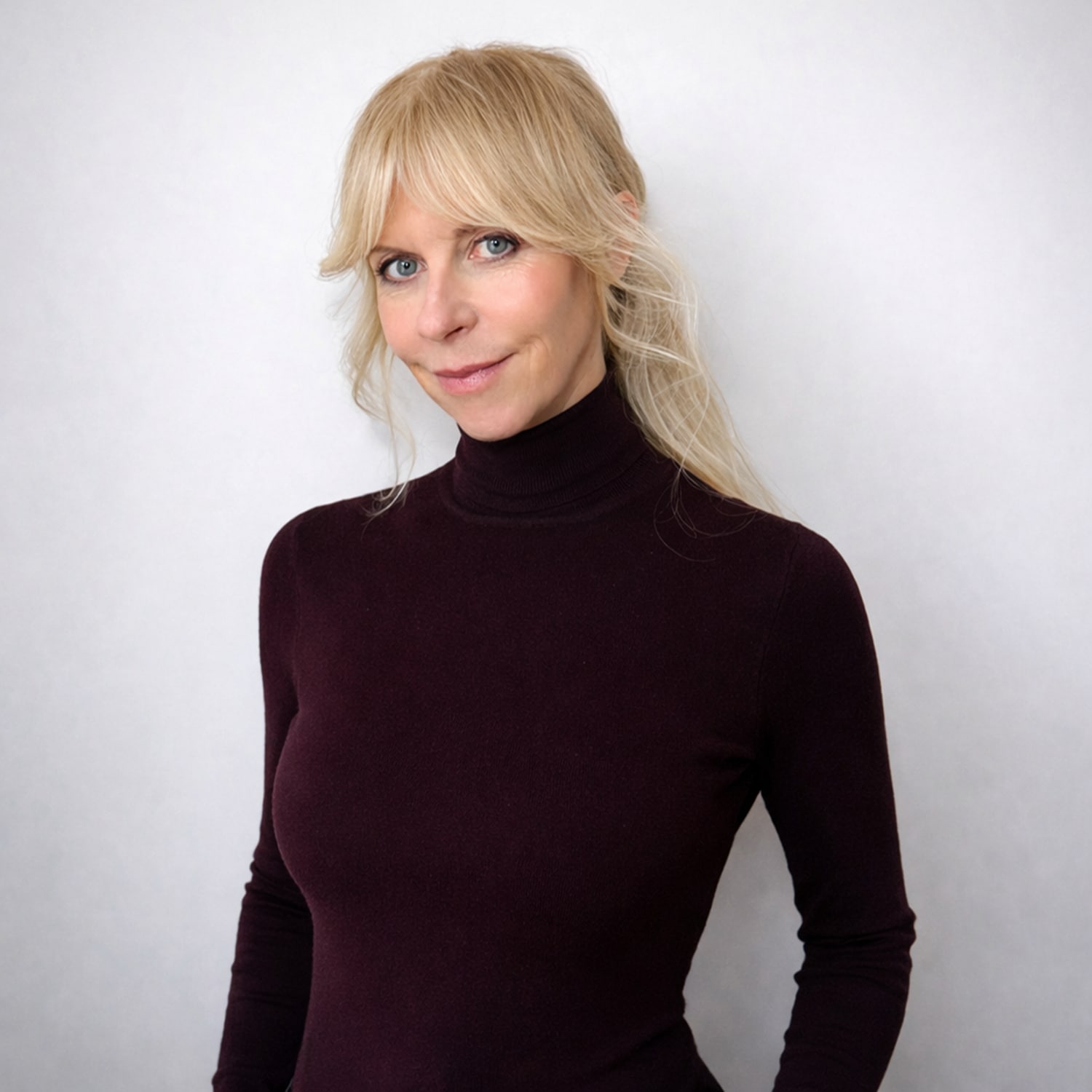 Porträt einer Frau mit blonden, schulterlangen Haaren und blauen Augen, die einen dunkelroten Rollkragenpullover trägt.