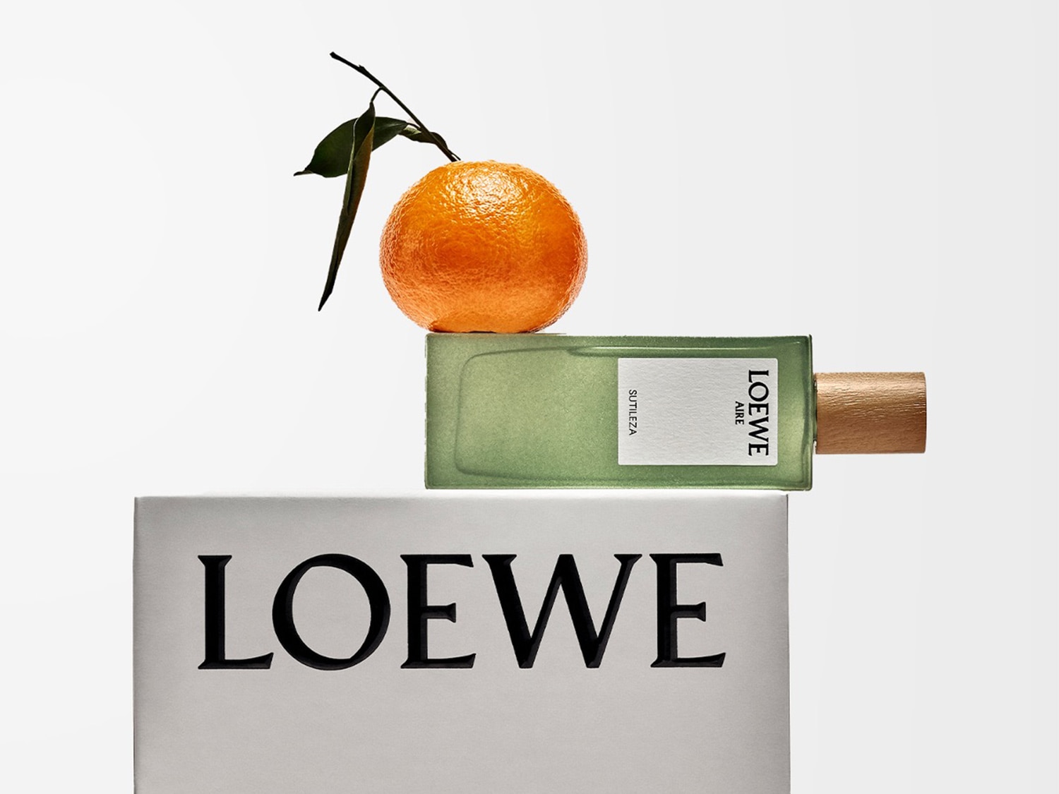Fragancia Loewe Aire Sutileza en frasco verde claro, coronada con una naranja fresca y vibrante, sobre una base con el logo de Loewe.