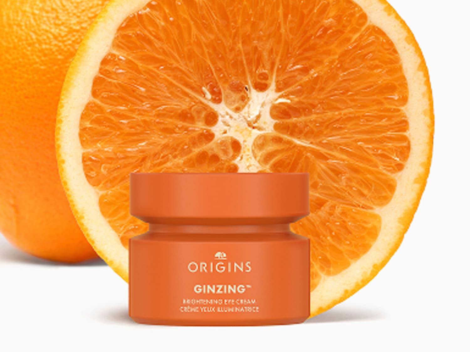 Een oranje potje Origins Ginzing Brightening oogcrème staat voor een halve sinaasappel, wat de verhelderende eigenschappen van de crème benadrukt.