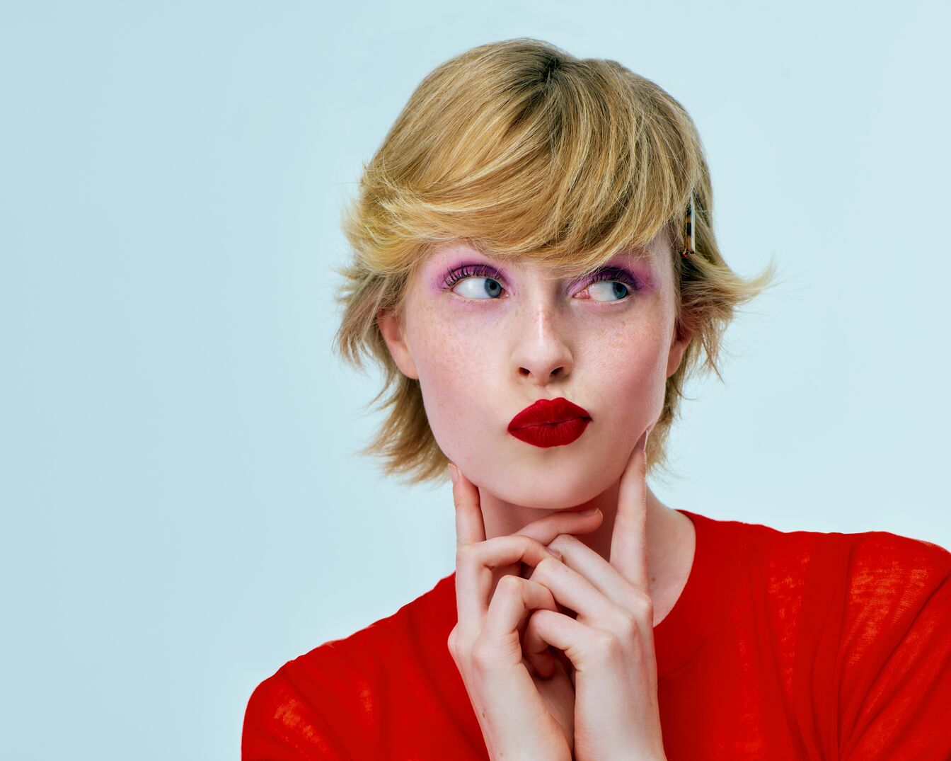 Portret kobiety z krótkimi blond włosami, różowym makijażem oczu i czerwoną szminką, ubranej w czerwoną koszulkę, zamyślonej i dotykającej palcami brody.