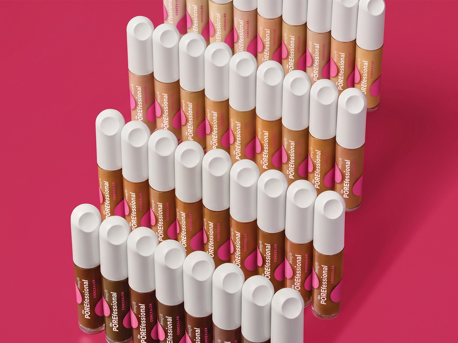 Een reeks Benefit POREfessional concealers in verschillende huidtinten, van licht tot donkerbruin, staan rechtop met witte doppen op een levendige roze achtergrond.