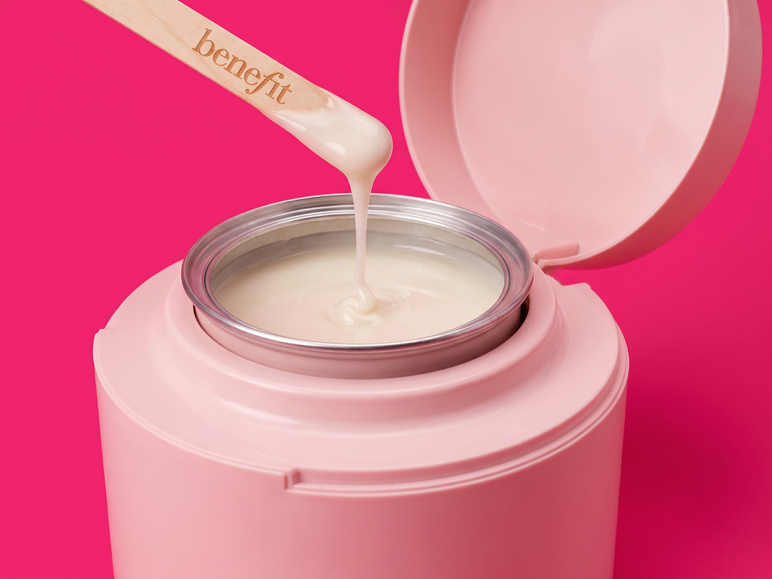 Een houten spatel met het merk "Benefit" schept romige, lichtbeige wax uit een roze waxverwarmer, tegen een levendige roze achtergrond.