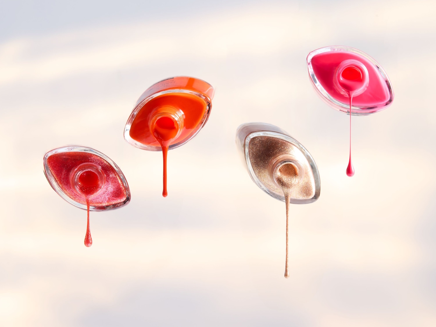 Vier Nagellackflaschen schweben auf einem hellen Hintergrund, wobei jeder Nagellack in den Farben Rot, Orange, Gold und Pink ausläuft.