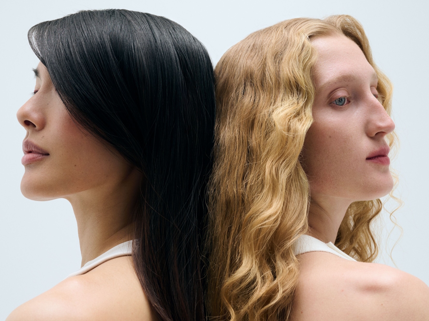 Zwei Frauen mit unterschiedlichen Haarfarben, eine mit glattem schwarzem Haar und die andere mit welligem blondem Haar, stehen Rücken an Rücken und präsentieren ihre gesunden Haare.