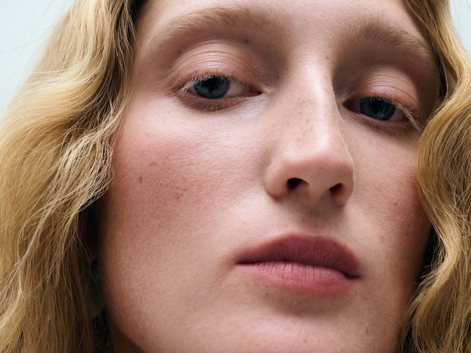 Eine Nahaufnahme zeigt das Gesicht einer Frau mit blondem, gewelltem Haar, blauen Augen und einem natürlichen Make-up-Look, der ihre Haut mit Sommersprossen und rosafarbenen Lippen betont.