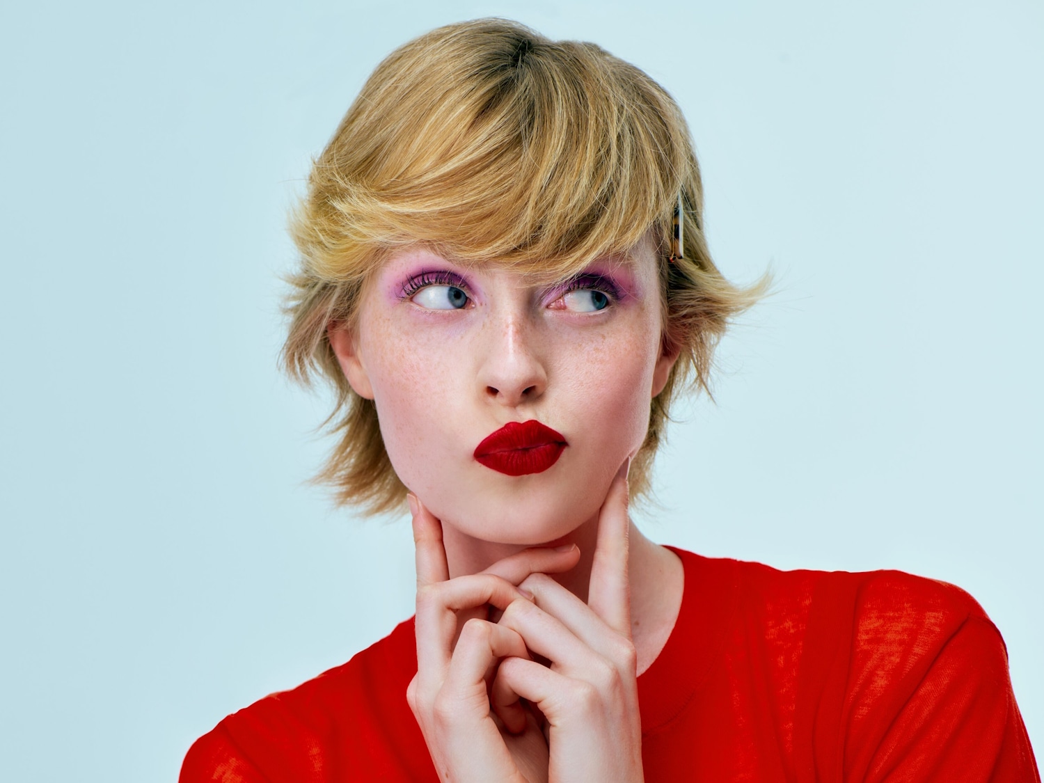 Eine Frau mit blonden, gewellten Haaren, rotem Lippenstift und einem roten Oberteil blickt mit einem nachdenklichen Ausdruck zur Seite.