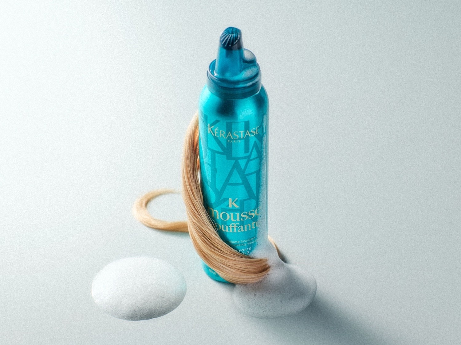 Blaue Kérastase-Flasche mit Mousse, umhüllt von einer blonden Haarsträhne, die die Textur und Anwendung des Produkts hervorhebt.