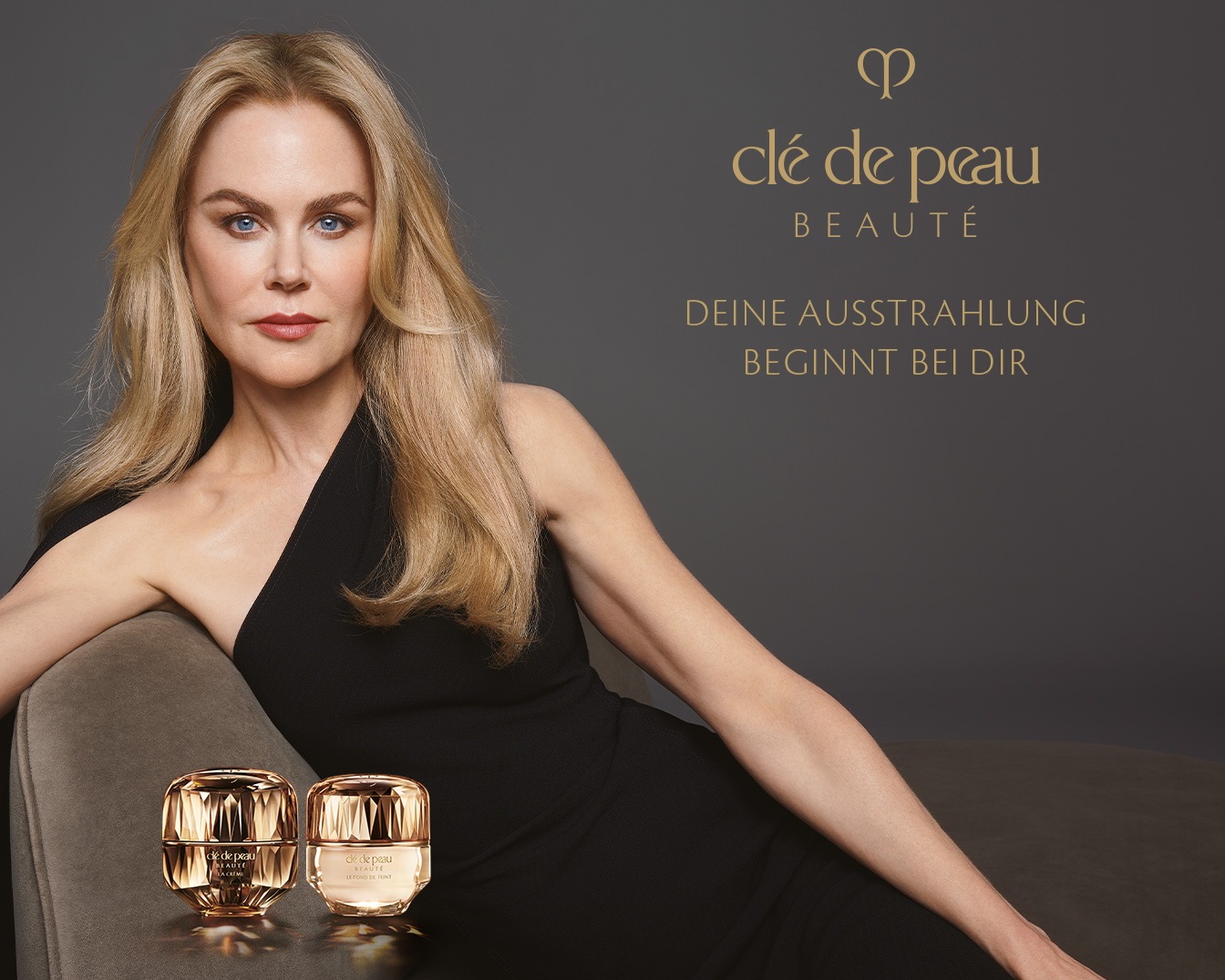 Eine blonde Frau mit einem schwarzen Kleid posiert neben zwei goldenen Tiegeln von Clé de Peau Beauté, darüber steht der Slogan: Deine Ausstrahlung beginnt bei dir.