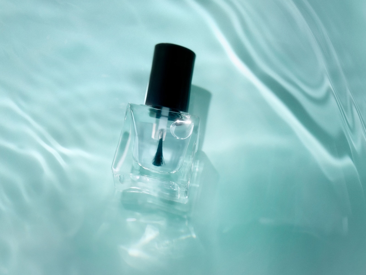 Heldere nagellak in een transparant flesje met een zwarte dop, liggend op een zachtblauwe achtergrond die het effect van water simuleert.