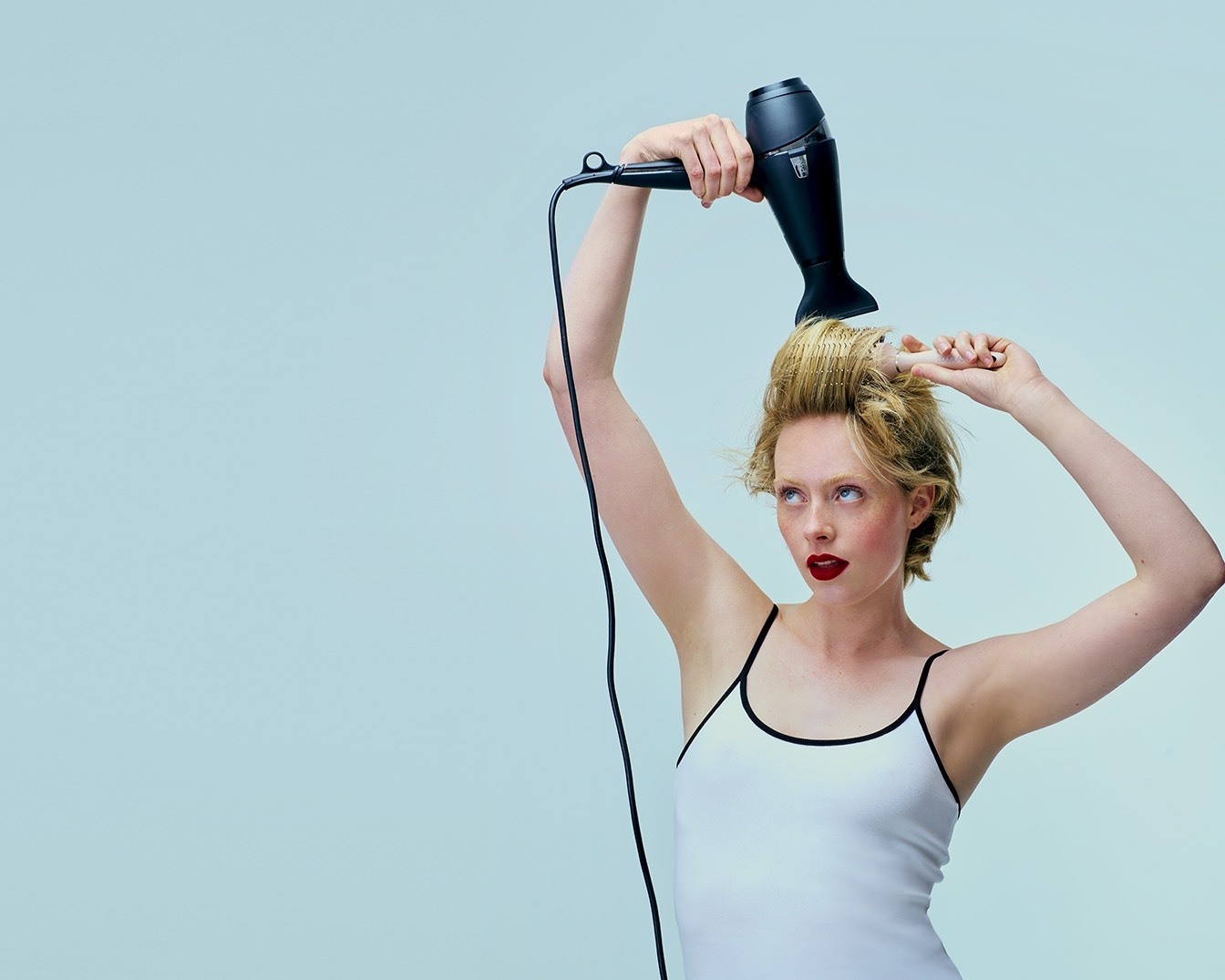Une femme aux cheveux blonds coiffe ses cheveux courts avec une brosse ronde et un sèche-cheveux noir sur fond bleu clair.