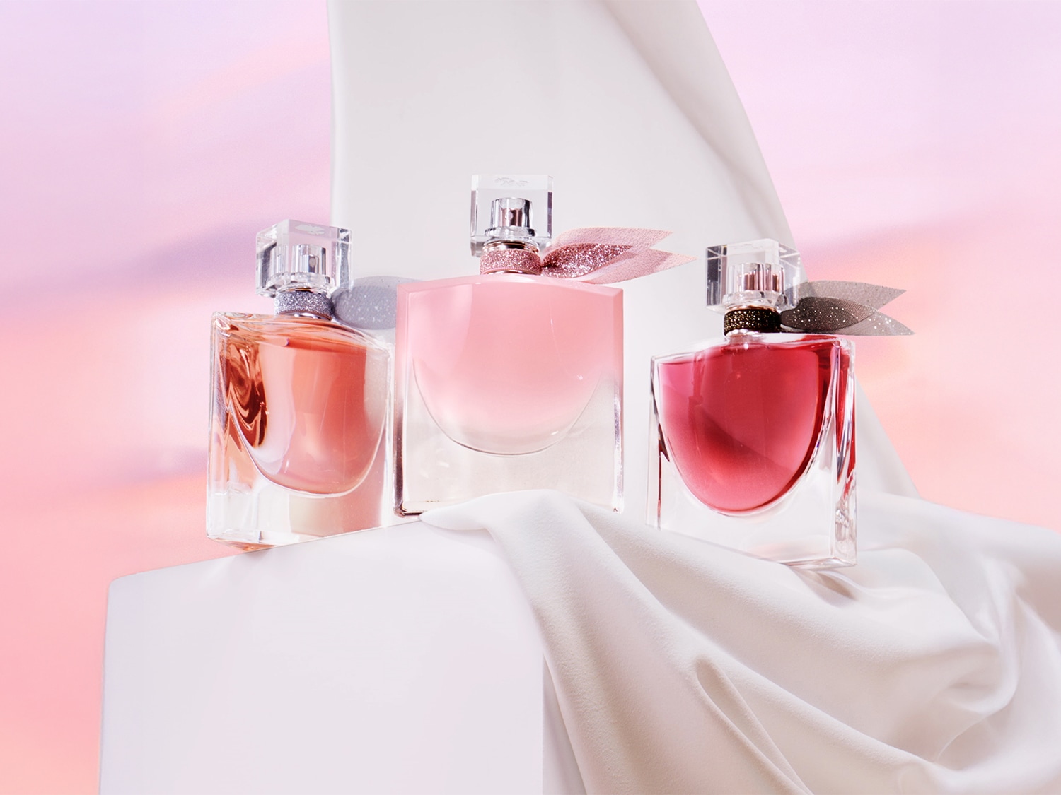 Drie La Vie Est Belle-parfums staan op een witte doek, elk met een andere tint roze en een sprankelende strik.