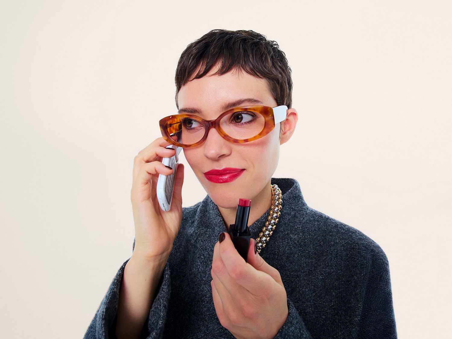 Une femme aux cheveux courts, vêtue d'un pull gris et d'un collier doré, porte des lunettes à monture écaille de tortue et blanche, parle au téléphone tout en tenant un rouge à lèvres rose vif.