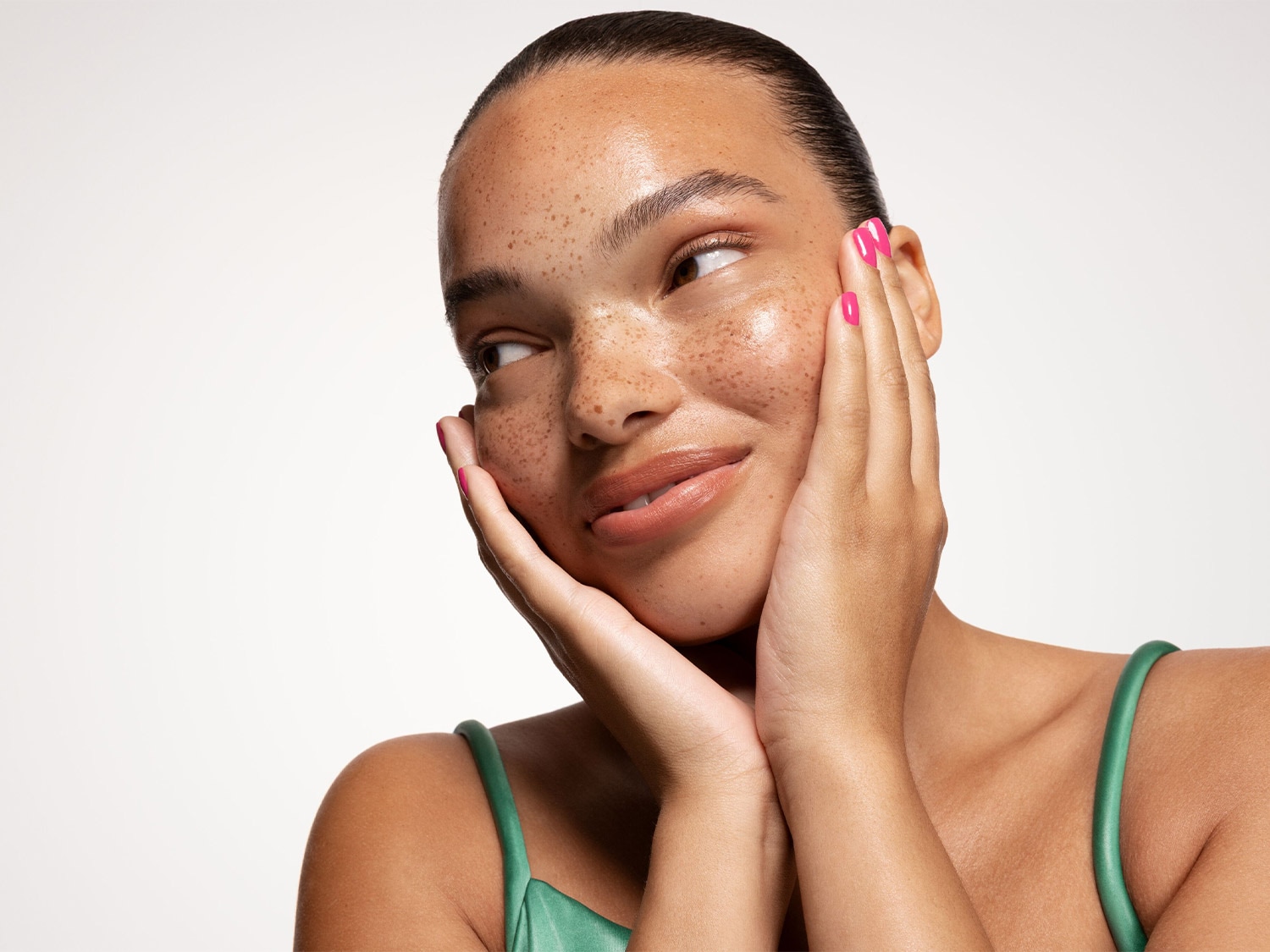Une femme souriante, la peau éclatante et parsemée de taches de rousseur, touche délicatement son visage des deux mains, arborant une manucure rose vif.