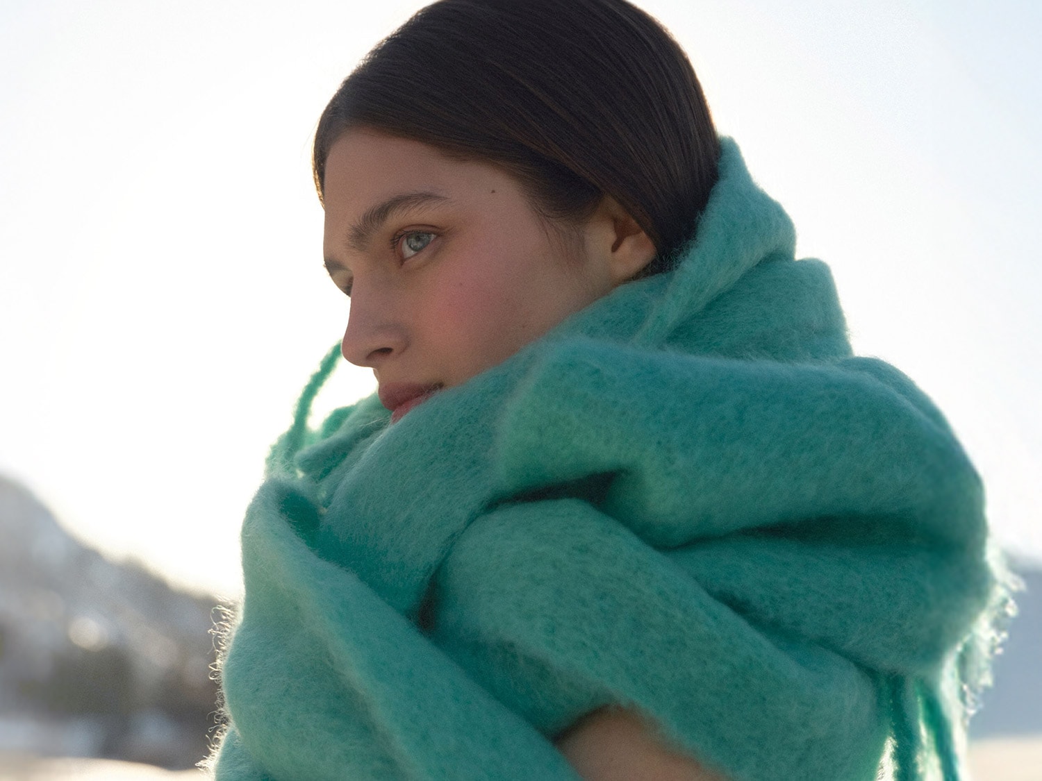 Une femme enveloppée dans une écharpe turquoise douce, regardant au loin avec une expression sereine.