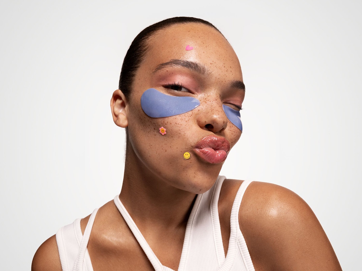 Une femme avec des taches de rousseur et des patchs bleus sous les yeux, agrémentée de petits autocollants ludiques en forme de cœur, de fleur et de smiley.