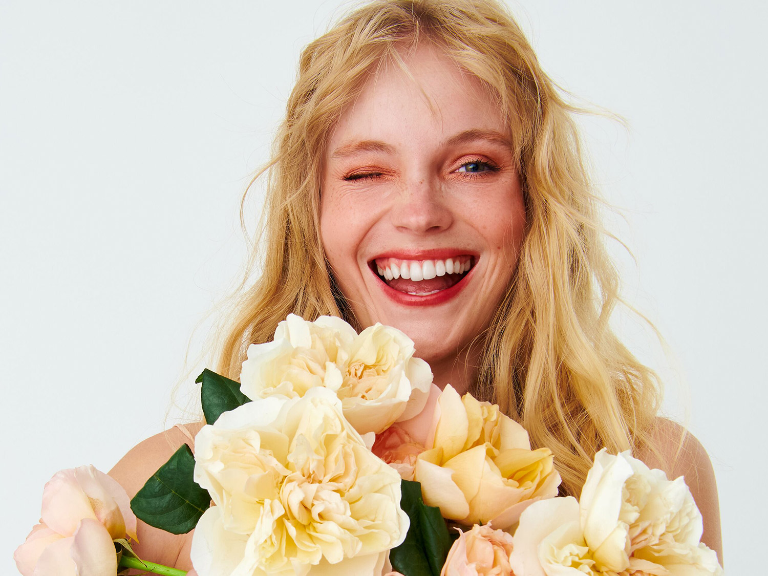 Une femme blonde souriante, un œil fermé, tient un bouquet de roses jaunes et pêche, rayonnant de joie et de fraîcheur.