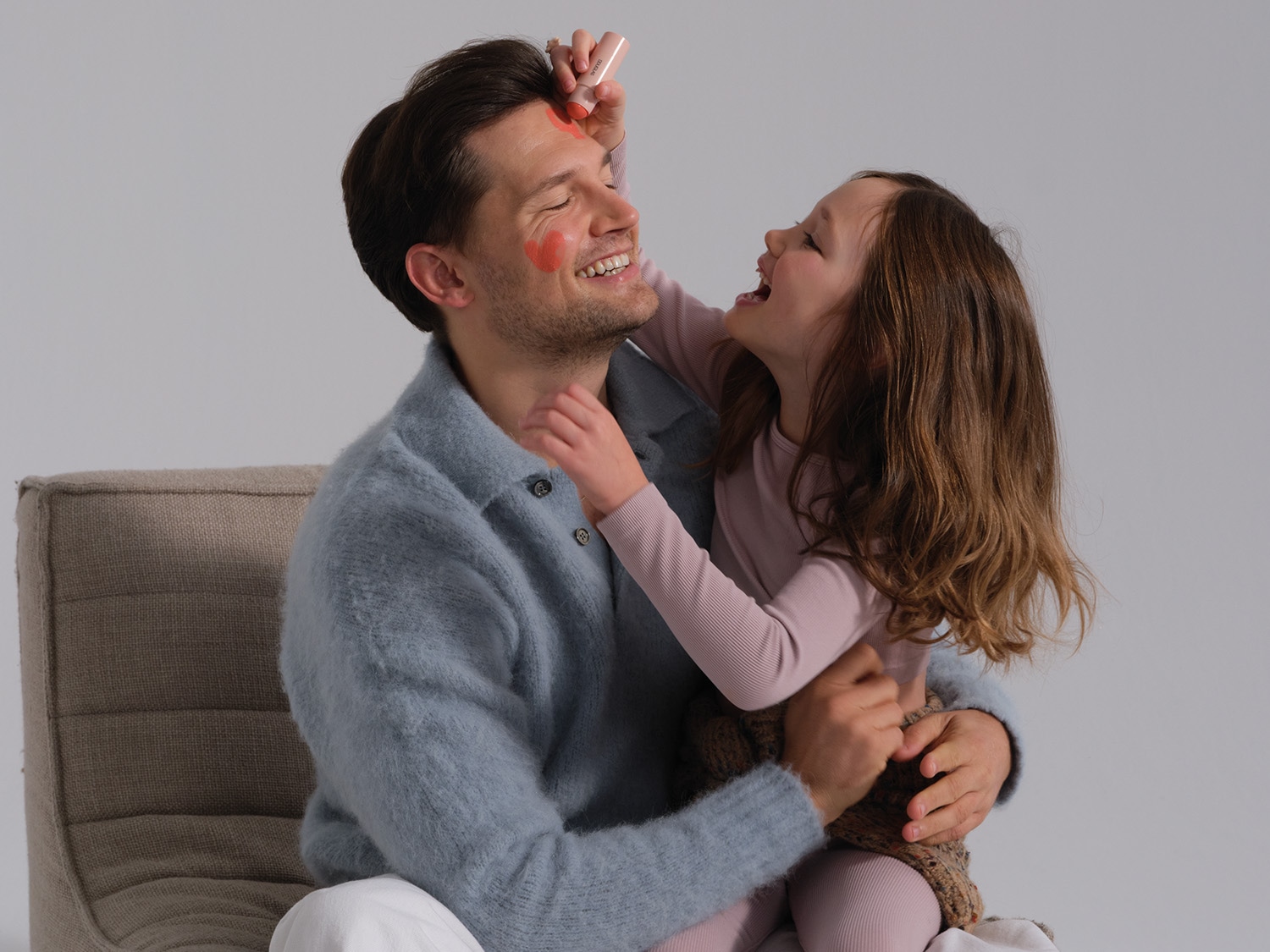 Un père souriant, assis, reçoit des cœurs rouges dessinés sur son visage par sa fille, qui rit en le serrant dans ses bras.