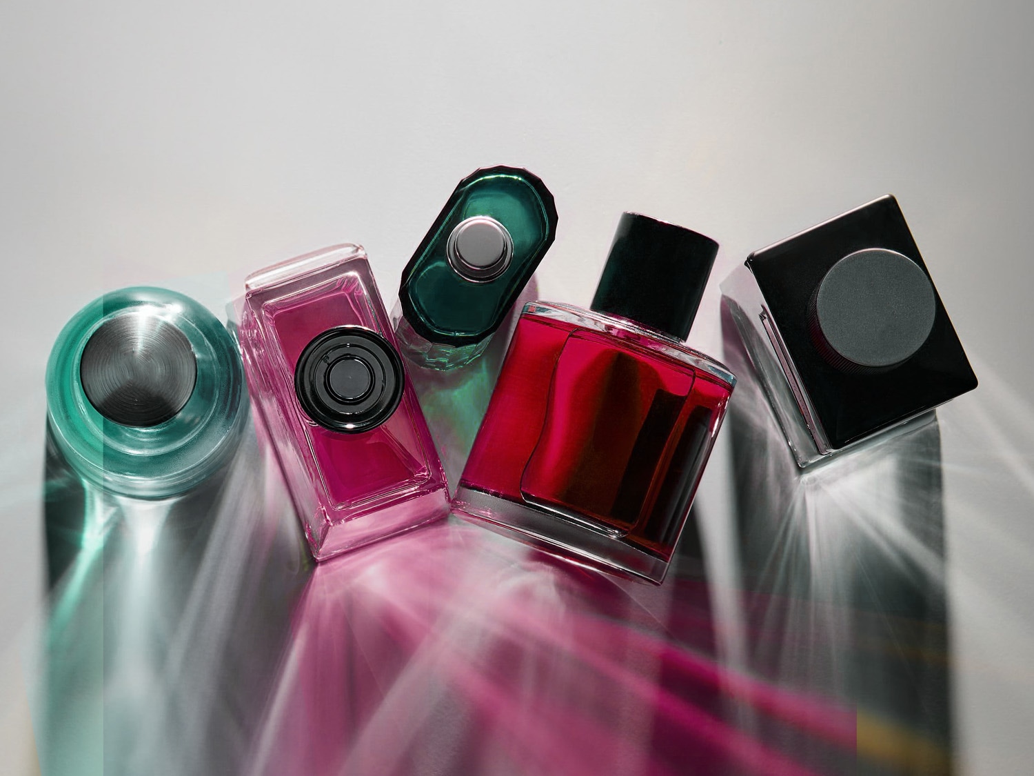 Cinq flacons de parfum aux couleurs vives, dont un vert sarcelle, un rose vif, un vert foncé, un rouge foncé et un noir, sont disposés sur une surface blanche, créant des reflets colorés.