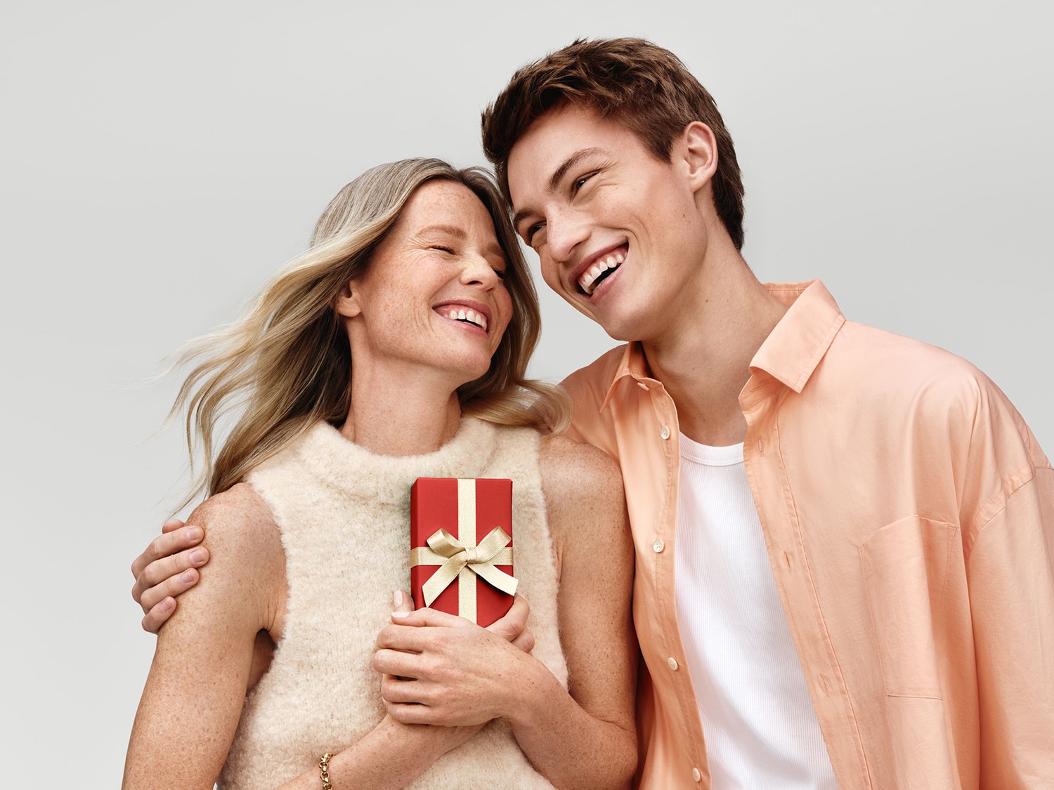 Un jeune homme souriant en chemise pêche enlaçant une femme blonde tenant un cadeau rouge orné d'un ruban doré, tous deux rayonnant de bonheur.