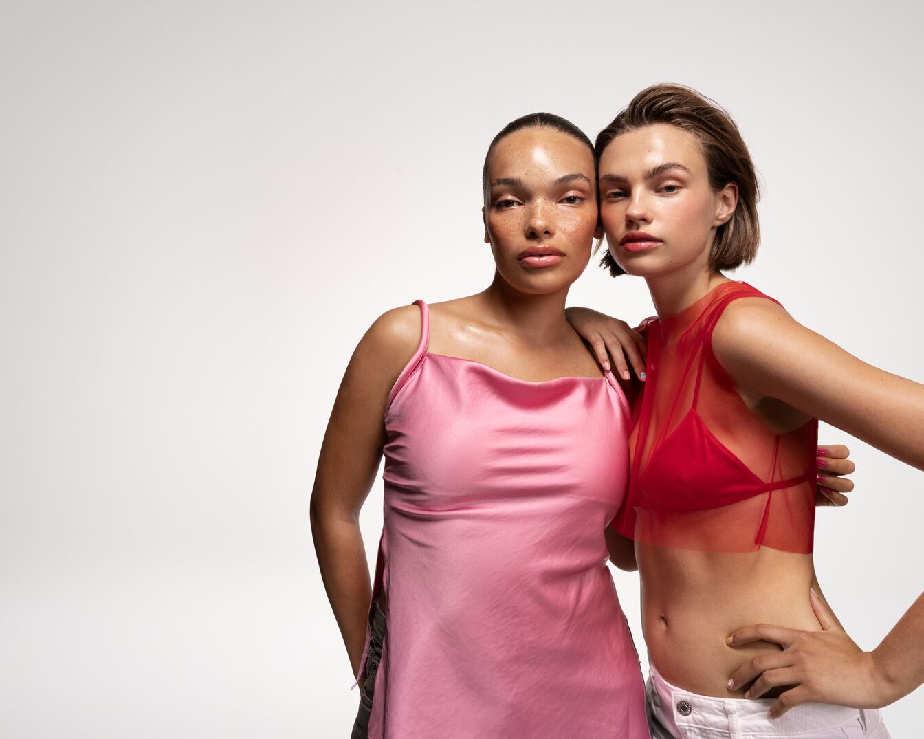 Dos modelos posan juntas, una con un vestido rosa satinado y la otra con una blusa roja transparente y pantalones blancos, ambas con maquillaje natural.