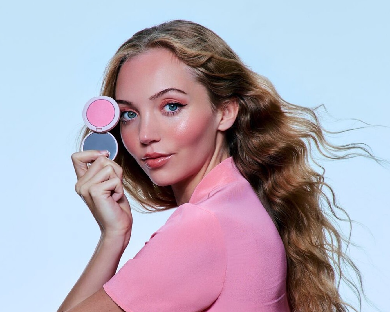 Eine Frau mit blonden, gewellten Haaren und einem rosa Blazer hält einen runden, rosa Rouge-Stick mit Spiegel in der Hand.
