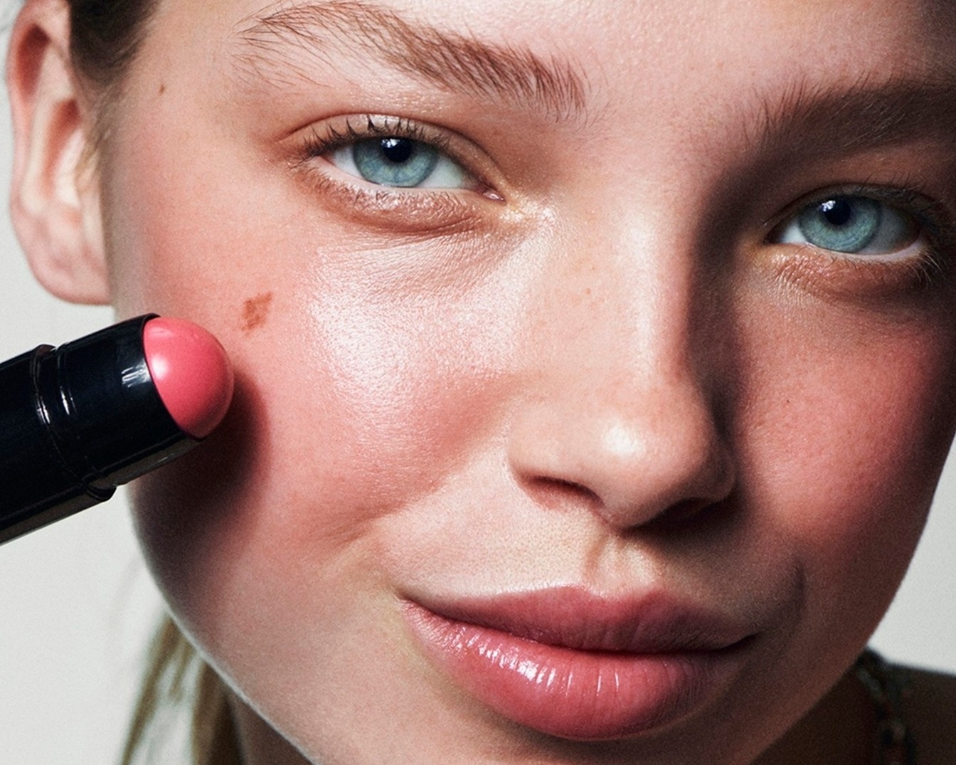 Egy fiatal nő arcának közeli képe, amint egy rózsaszín krémes pirosítót visz fel az arcára egy fekete rúdból, kiemelve a természetes ragyogást.