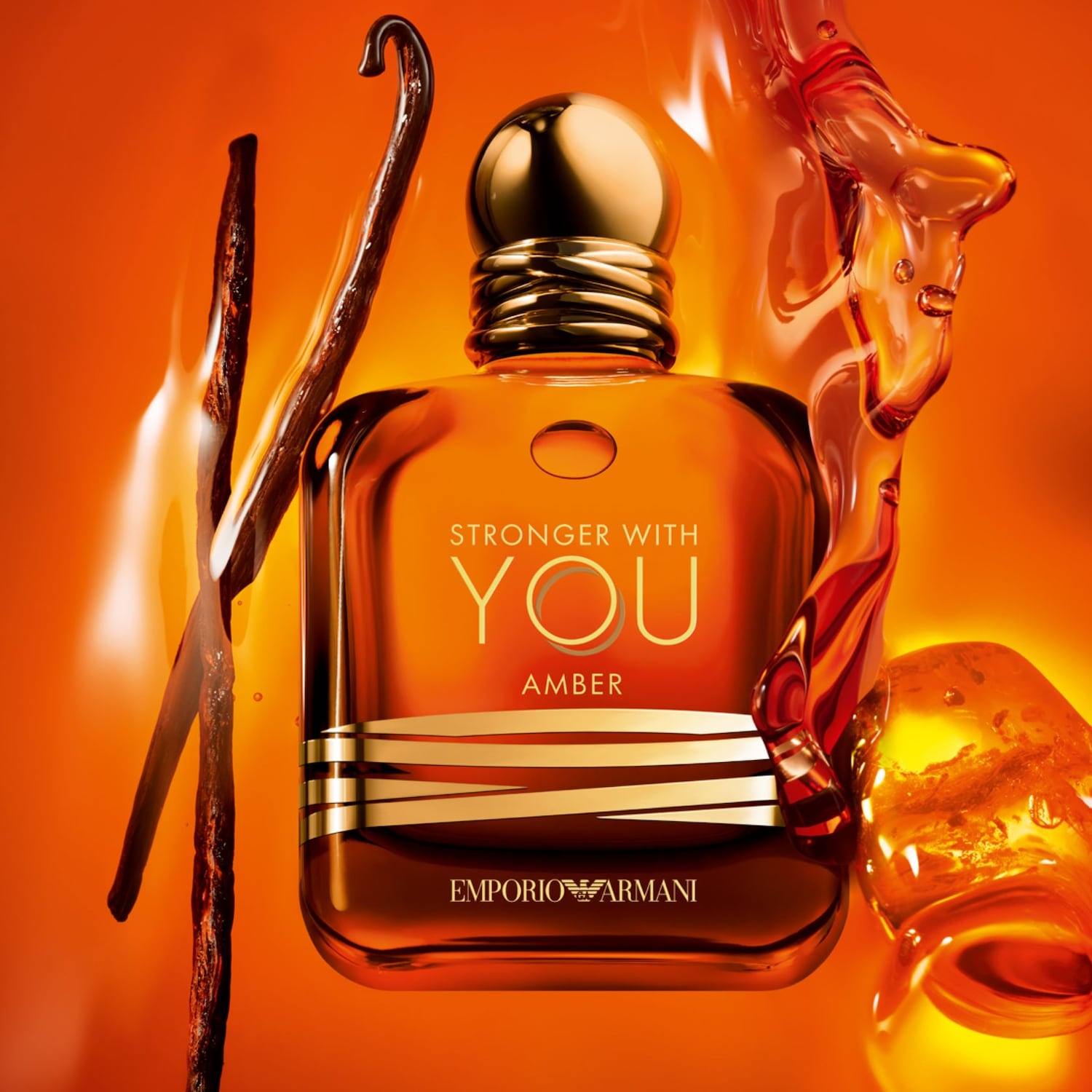 Narancssárga háttér előtt az Emporio Armani Stronger With You Amber parfümös üvege látható, mellette vaníliarúd és borostyánszínű folyadék.