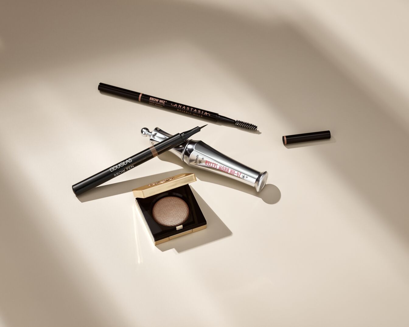 Verschiedene Augenbrauenprodukte, darunter ein Douglas Brow Pen, ein Anastasia Brow Wiz, ein Benefit 24-HR Brow Setter und ein goldener Lidschatten, liegen auf einem hellen Hintergrund.