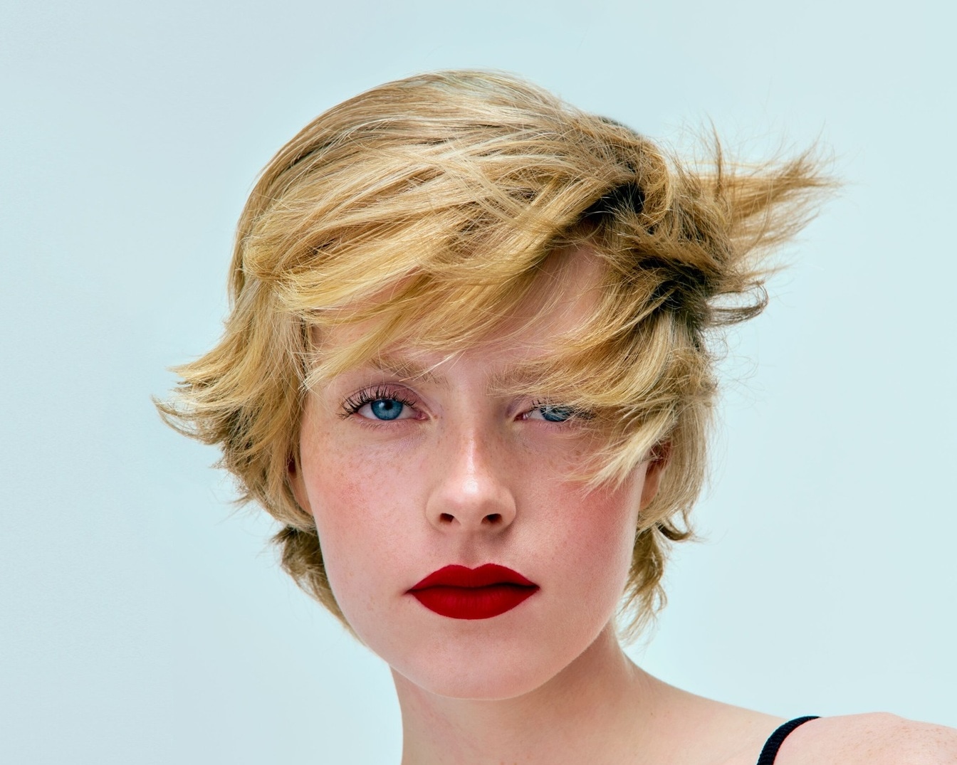 Nahaufnahme eines jungen Models mit blonden, gewellten, kinnlangen Haaren, blauen Augen und roten Lippen.