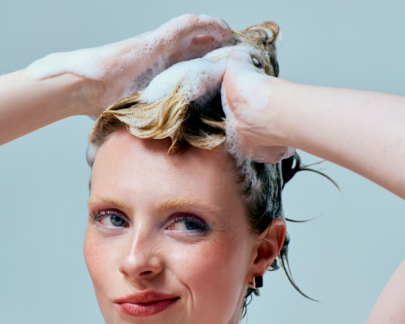 Nahaufnahme einer Frau mit hellblondem, gewelltem Haar, das mit weißem Schaum bedeckt ist, während sie es mit beiden Händen einshampooniert.
