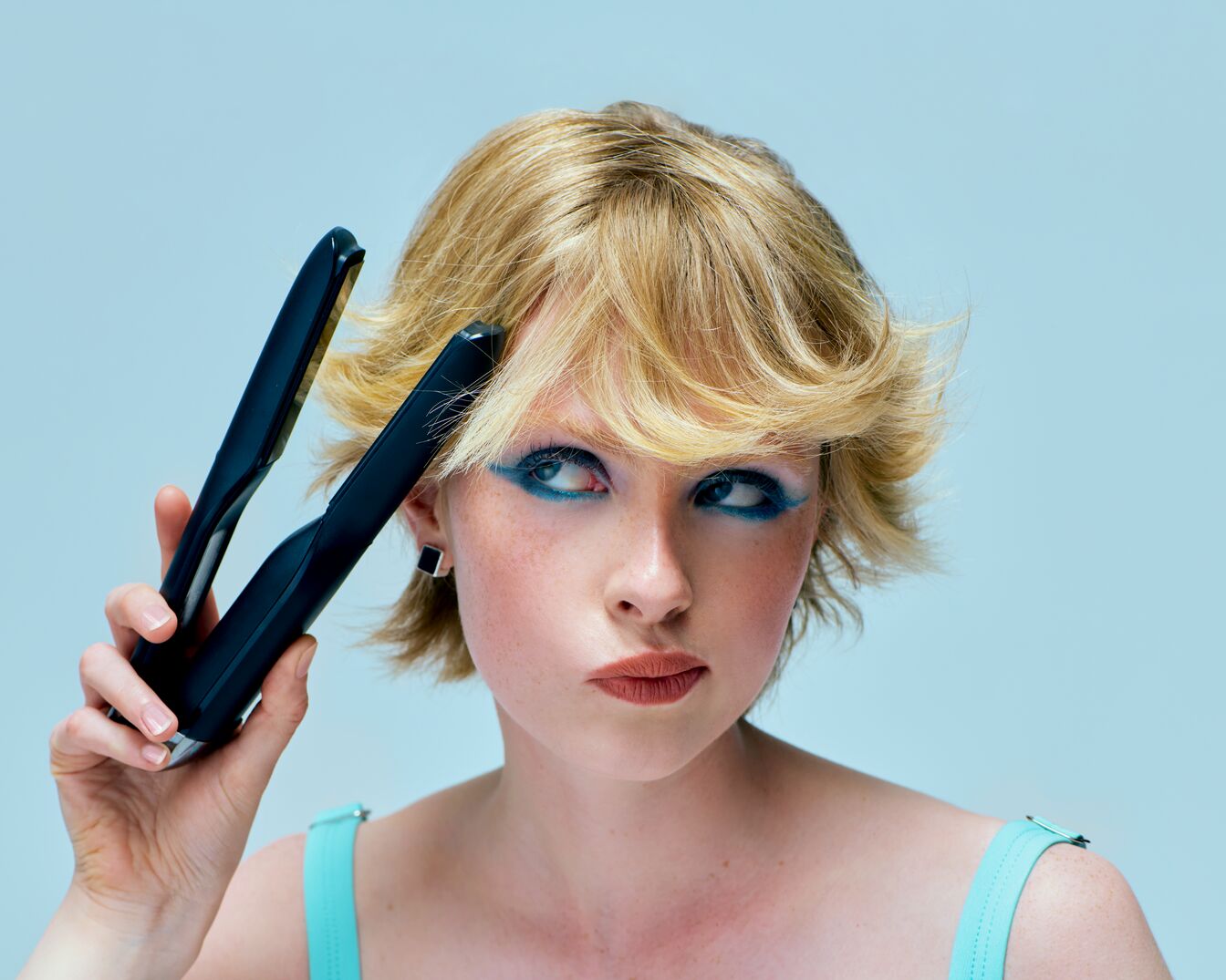 Eine Frau mit blonden, gewellten Haaren und blauem Lidschatten hält ein schwarzes Glätteisen in der Hand und schaut zur Seite.