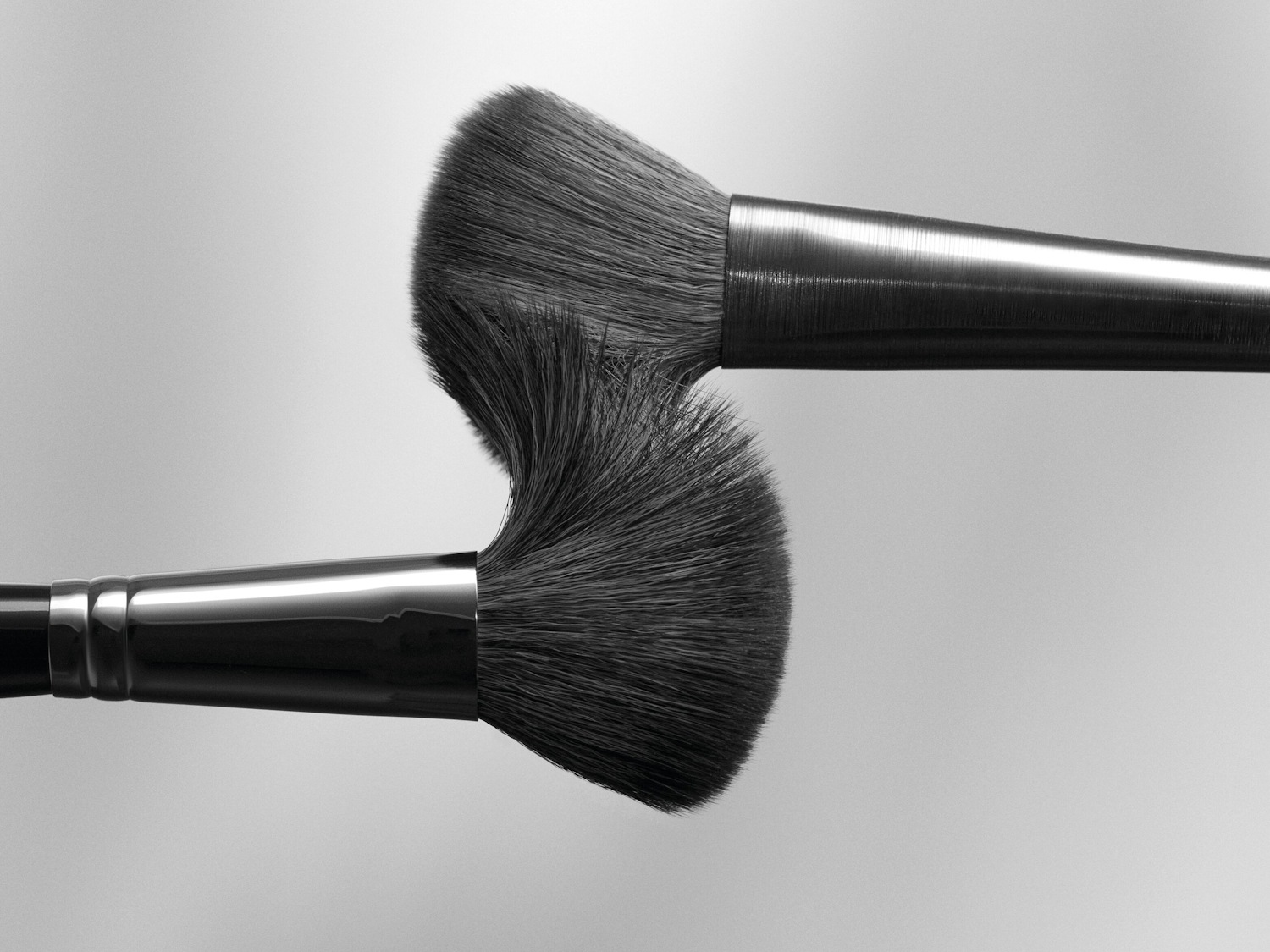 Zwei Make-up-Pinsel mit dicken, weichen Borsten und glänzenden, metallischen Griffen kreuzen sich vor einem neutralen Hintergrund.
