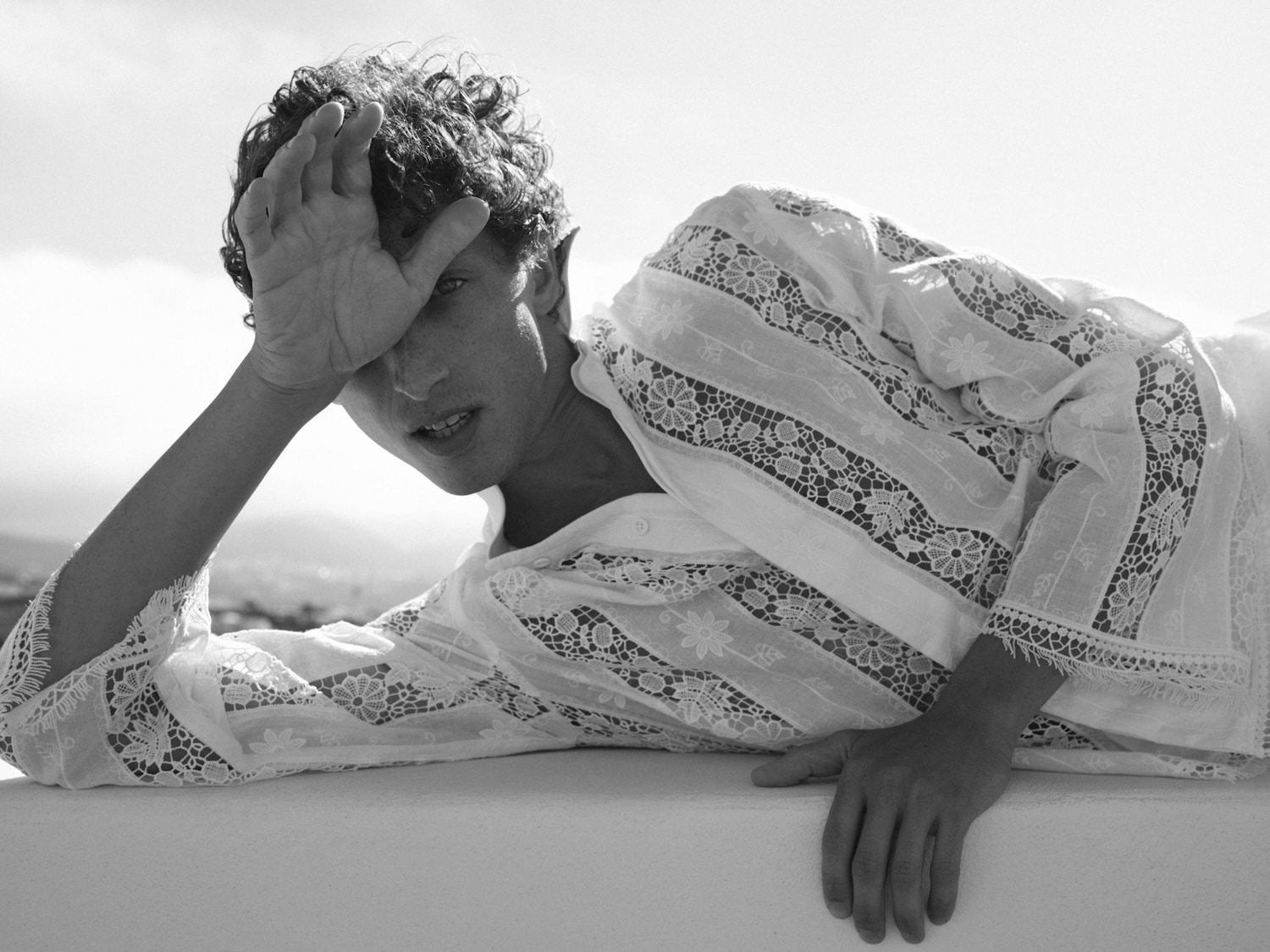 En una foto en blanco y negro, un modelo con cabello rizado y una camisa blanca de encaje yace sobre una superficie blanca, con la mano en la frente, mirando a la cámara.