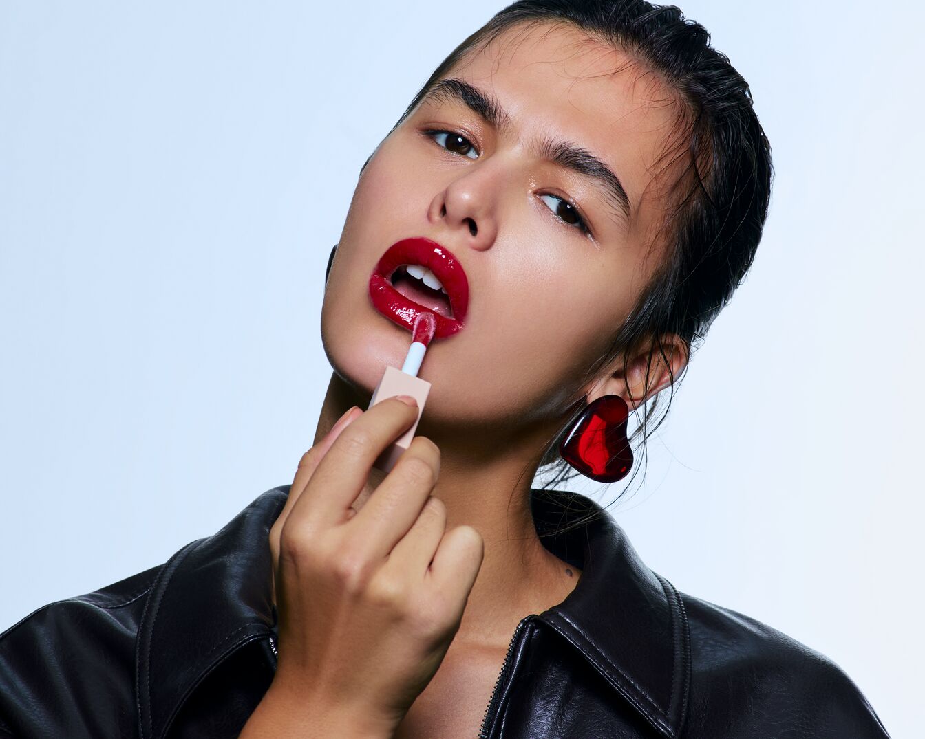 Eine Frau mit glänzenden roten Lippen trägt eine schwarze Lederjacke und herzförmige Ohrringe, während sie mit einem Pinsel roten Lipgloss aufträgt.