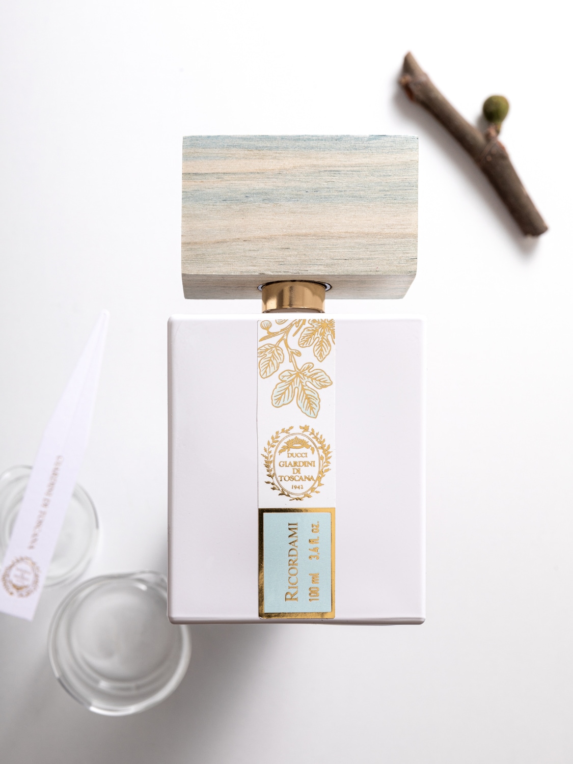 De Ricordami parfumfles van 100 ml heeft een witte, vierkante vorm met een lichtblauwe en gouden label, en een houten dop met een gouden hals, naast een takje en twee glazen.