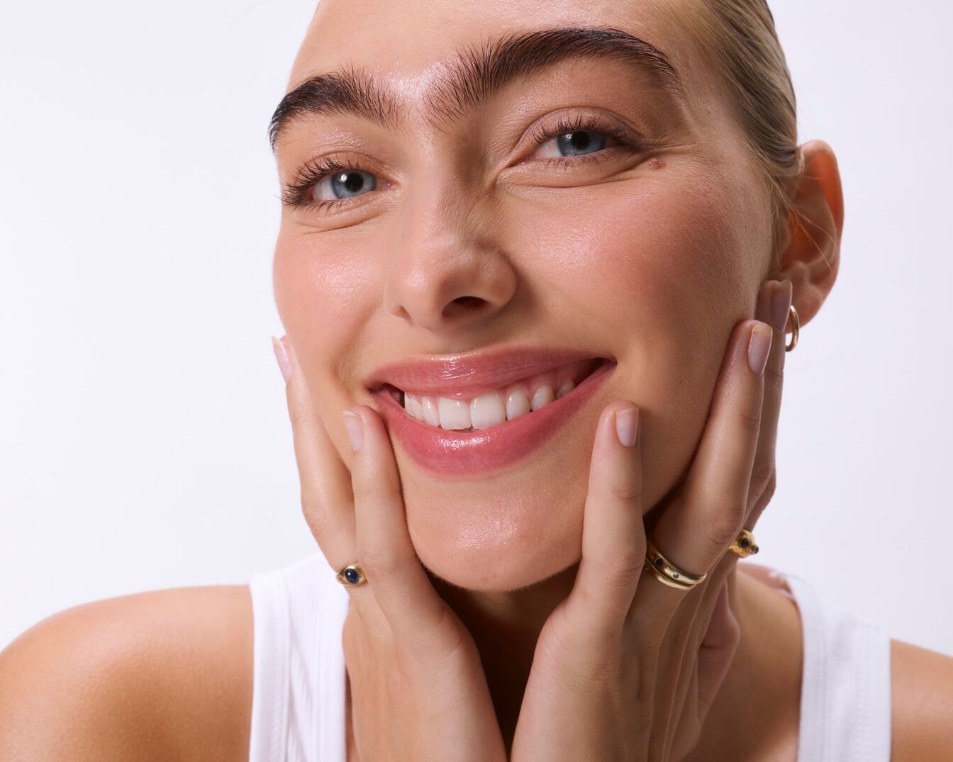 Eine junge Frau mit strahlendem Lächeln und glänzenden Lippen, die ihre Wangen mit den Händen berührt, trägt einen goldenen Ohrring und goldene Ringe an ihren Fingern.
